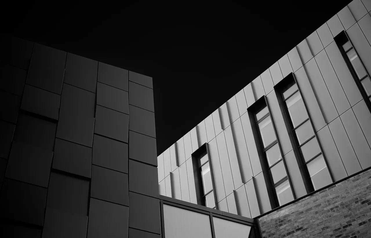 Two modern building facades meeting at a corner against a black sky, one clad in staggered panels and one in vertical ribbing