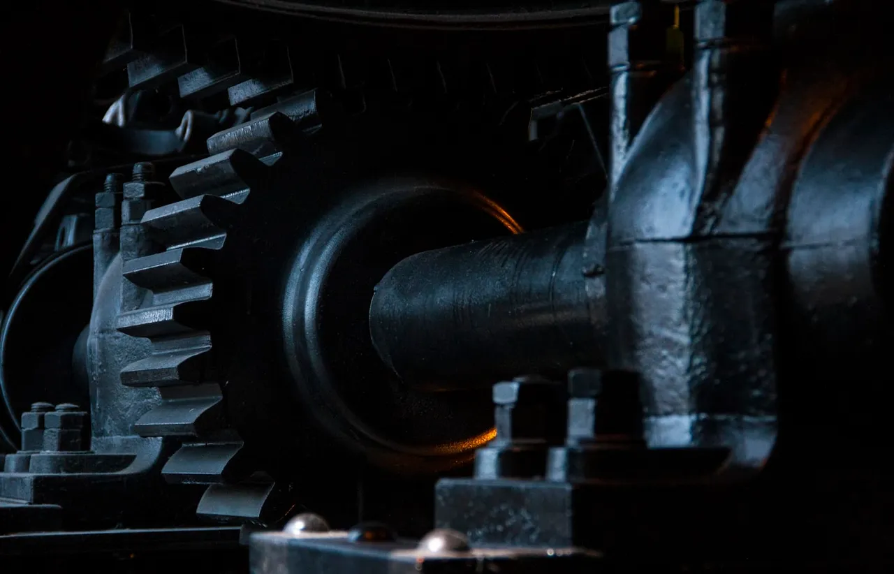 Close up of a large black cast iron cogwheel meshing with bolted machinery and a heavy drive shaft in low side light