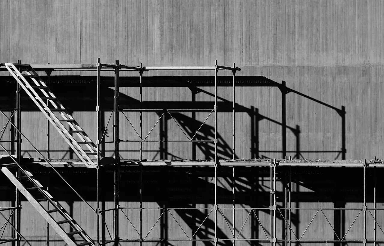 Black and white multi level metal scaffolding with diagonal stair flights and cross braces casting angular shadows on a concrete wall